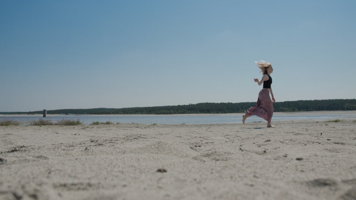 A young woman in a black top runs on a sandy beach, enjoying a sunny day.